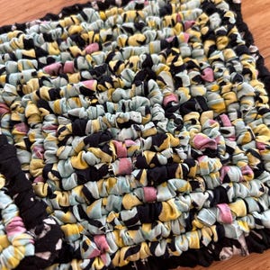 May include: A square fabric potholder constructed from braided strips of cloth in a multi-colored pattern. The textile features a repeating color palette of black, pale blue, yellow, and soft pink, woven into a dense, textured, and thick surface.