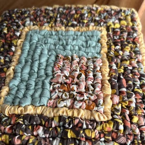 May include: A square, handmade textile piece with a patchwork design. The center is a light blue square, surrounded by a yellow border, and then a patchwork of brown, yellow, and floral fabrics. The outer edges are a mix of brown, yellow, and floral fabrics.