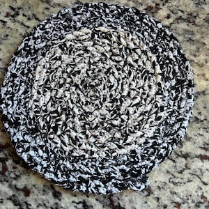 May include: A round, handmade trivet with a black and white floral pattern. The trivet is made of tightly woven fabric strips, creating a textured surface. The design features a spiral pattern, with the black and white elements alternating.