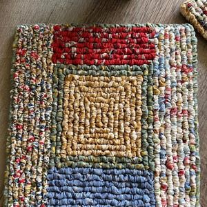 May include: A handmade, square fabric trivet with a patchwork design. The trivet features a central square of golden yellow fabric surrounded by red, blue, and floral patterned strips. The edges are finished with a green border.