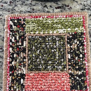 May include: A square, handmade rag rug with a patchwork design. The rug features sections of black, green, red, and white fabric strips, bordered by a tan fabric. The edges are finished with a red and white striped trim.