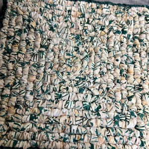 May include: A rectangular, handmade rug with a woven texture. The rug features a cream-colored base with a repeating pattern of green leaf-like shapes. The edges are trimmed with a dark green border. The rug is approximately 12 inches long and 8 inches wide.