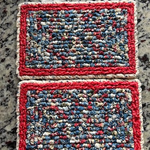 May include: Two rectangular, handmade placemats. The center of each mat is a patchwork of blue, red, and white fabric strips. A red border surrounds the patchwork, and a cream-colored, ruffled edge finishes the design. The mats are likely for kitchen or dining use.