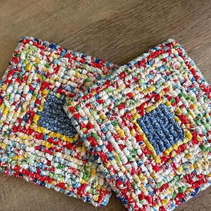 May include: Two square, handmade trivets. The trivets are made of woven fabric strips in a patchwork design. The colors include red, blue, yellow, and white. The center of each trivet is a solid blue square.