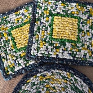 May include: Handmade, colorful fabric trivets and coasters. The trivets are square with a yellow center, green border, and white, green, and yellow accents. The coasters are round with similar colors and patterns. The edges are a dark blue.