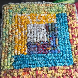 May include: A square fabric mat constructed from concentric rows of tightly gathered, multi-colored textile strips. The design features a central white square surrounded by layers of yellow, blue, purple, green, and pink patterned fabric pieces.