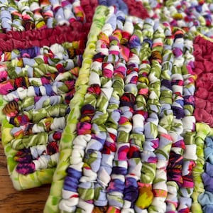 May include: Close-up of colorful, braided fabric placemats. The placemats feature a patchwork design with a variety of floral and solid fabric scraps in shades of green, pink, purple, and blue. The edges are finished with a green border.
