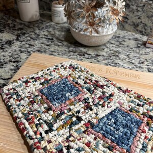 May include: Two square potholders crafted from woven fabric strips in a multicolored pattern. Each potholder features a central square of blue floral fabric surrounded by concentric rows of white, red, yellow, and dark blue patterned textile strips.