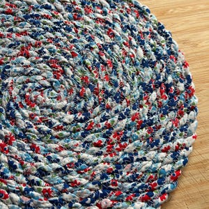 May include: A circular coaster or trivet constructed from coiled fabric strips featuring a floral pattern in shades of red, blue, and white. The fabric coils are joined together with visible white zigzag stitching, creating a textured surface.
