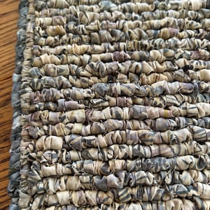 May include: Close-up of a handmade, rectangular rug with a textured surface. The rug is made of tightly woven fabric strips in shades of beige, gray, and muted purple. The fabric strips have a subtle pattern, creating a unique, artisanal look.