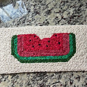 May include: A rectangular rug with a watermelon slice design. The watermelon has a red center with black seeds, a green rind, and a white border. The rug has a woven texture and a neutral background.