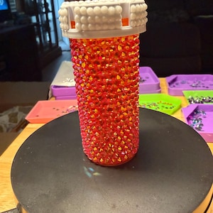 May include: A bedazzled pill bottle, covered in red and orange rhinestones. The bottle has a white lid with a textured design. The bottle is displayed on a black rotating surface, with colorful trays in the background.