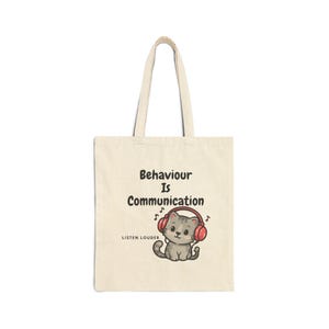 Può includere: Borsa tote in tela beige con testo nero "Behaviour Is Communication" e "Listen Louder". Un gatto dei cartoni animati con cuffie rosse è sotto. La borsa ha due manici beige.