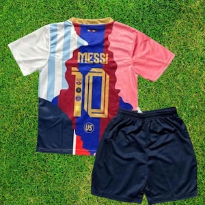 Pu&ograve; includere: Un completo da calcio composto da maglia e pantaloncini. La maglia &egrave; bianca, azzurra, rossa e blu navy, con il nome "MESSI" e il numero "10" sul davanti. I pantaloncini sono blu navy. Il completo &egrave; esposto su una superficie erbosa verde.
