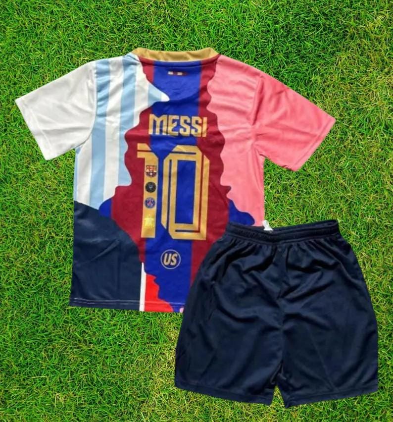 Pu&ograve; includere: Un completo da calcio composto da maglia e pantaloncini. La maglia &egrave; bianca, azzurra, rossa e blu navy, con il nome "MESSI" e il numero "10" sul davanti. I pantaloncini sono blu navy. Il completo &egrave; esposto su una superficie erbosa verde.