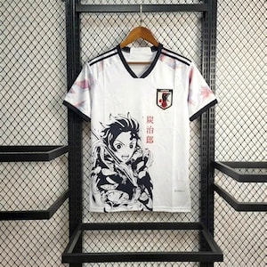 Puede incluir: Camiseta de fútbol blanca con detalles negros, cuello en V y el emblema de la bandera japonesa. Presenta un gráfico de personaje de anime en blanco y negro y texto japonés. La camiseta tiene rayas negras en los hombros.