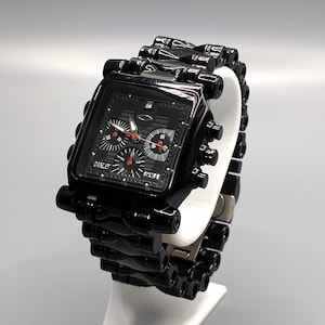 May include: A black Oakley wristwatch with a square face and a matching black band. The watch features red and white accents on the face and is displayed on a white stand against a gray background.