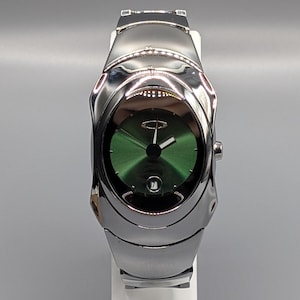 Könnte beinhalten: Eine silberfarbene Armbanduhr mit grünem Zifferblatt. Die Uhr hat ein schlankes, modernes Design mit einem Metallarmband und einem runden Zifferblatt. Die Uhr wird auf einem weißen Ständer vor grauem Hintergrund präsentiert.