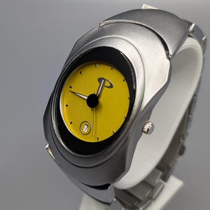 Pode incluir: Relógio de pulso prateado com mostrador amarelo. O relógio apresenta ponteiros pretos, uma janela de data e o logótipo Oakley. O relógio é exibido em um suporte branco sobre um fundo cinza.