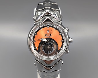 May include: A silver-toned Oakley wristwatch with an orange face and a smaller black dial. The watch has a metallic band and a date display. The watch is displayed on a white stand against a gray background.