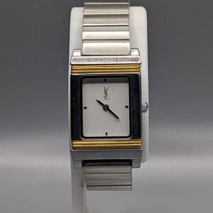 May include: A silver and gold-toned wristwatch with a rectangular white face. The watch features a silver band, gold accents, and a black frame around the face. The watch is displayed on a white stand.