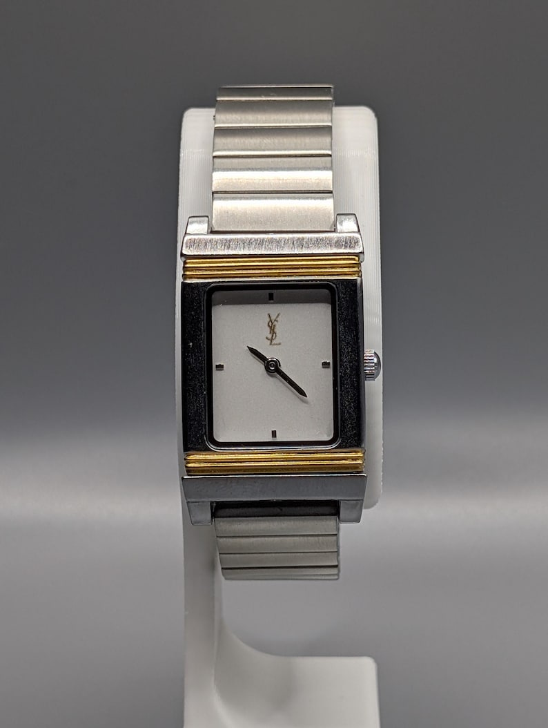 May include: A silver and gold-toned wristwatch with a rectangular white face. The watch features a silver band, gold accents, and a black frame around the face. The watch is displayed on a white stand.