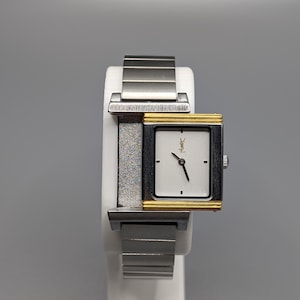 May include: A rectangular wristwatch with a silver band and a gold-toned bezel. The watch face is white with black hands and a small logo. The watch is displayed on a white stand against a gray background.