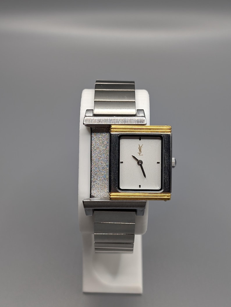 May include: A rectangular wristwatch with a silver band and a gold-toned bezel. The watch face is white with black hands and a small logo. The watch is displayed on a white stand against a gray background.