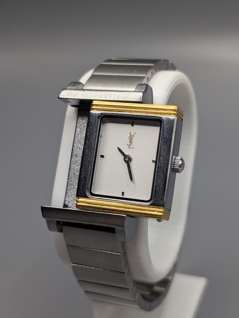 May include: A rectangular wristwatch with a silver-tone metal band and case. The watch face is white with black hands and a gold-tone border. The watch features the Yves Saint Laurent logo.