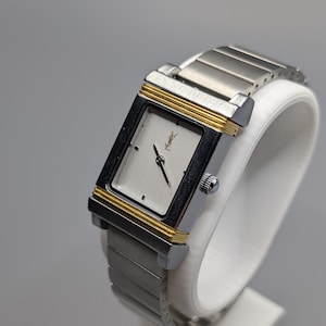 May include: A rectangular silver-toned wristwatch with gold-colored accents. The watch features a silver face with black hands and a small logo. The watch band is made of silver-toned metal links. The watch is displayed on a white stand.