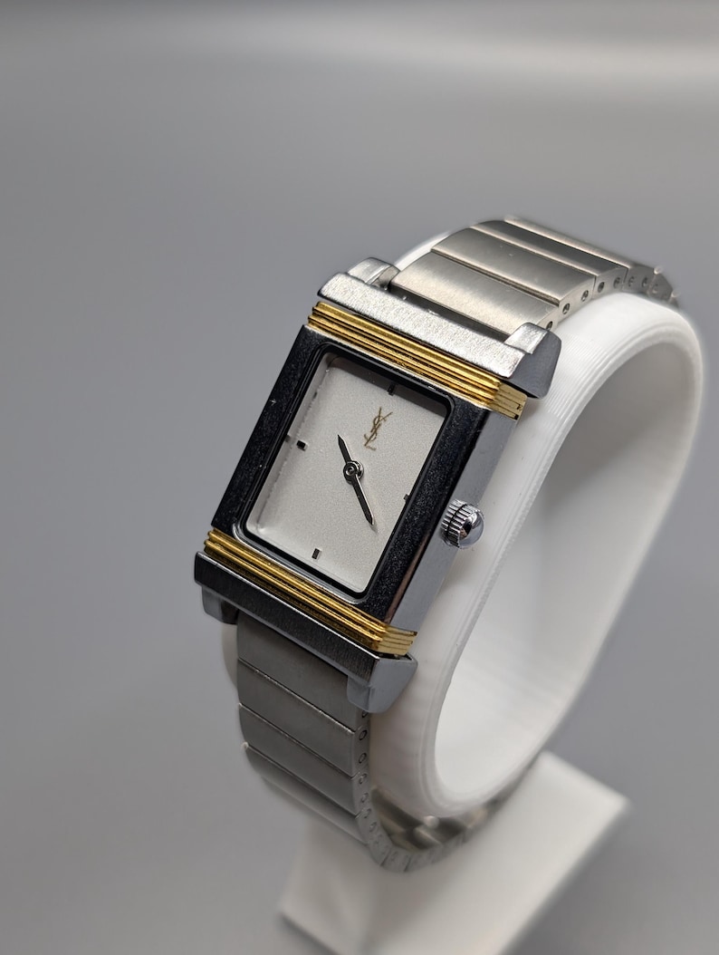 May include: A rectangular silver-toned wristwatch with gold-colored accents. The watch features a silver face with black hands and a small logo. The watch band is made of silver-toned metal links. The watch is displayed on a white stand.