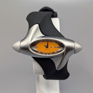 Könnte beinhalten: Eine silberne und schwarze Armbanduhr mit einem ovalen, orangefarbenen Zifferblatt. Die Uhr hat ein schwarzes Kautschukarmband und ein silbernes Gehäuse. Das Zifferblatt hat schwarze Zeiger und ein kleines Logo. Die Uhr wird auf einem weißen Ständer präsentiert.