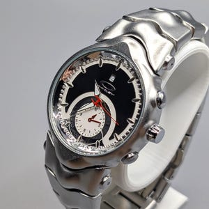 May include: A silver wristwatch with a black face and a white subdial. The watch has a red second hand and a silver link strap. The watch is displayed on a white stand against a grey background.