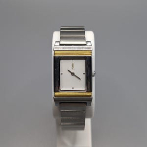 May include: A rectangular wristwatch with a silver band and a white face. The watch has a gold-colored frame around the face and a small logo at the top. The watch is displayed on a white stand.