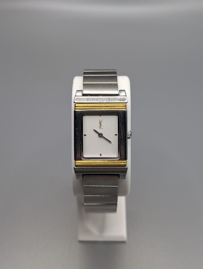 May include: A rectangular wristwatch with a silver band and a white face. The watch has a gold-colored frame around the face and a small logo at the top. The watch is displayed on a white stand.