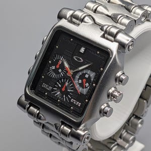 May include: A silver Oakley wristwatch with a black face and red detailing. The watch features a rectangular dial with multiple sub-dials and a silver metal strap. The watch is displayed on a white stand.
