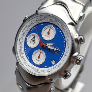 May include: A silver-toned wristwatch with a blue face and multiple sub-dials. The watch features a date display and a silver band. The face includes the text "LOCAL TIME" and city names around the perimeter.