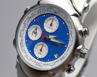 May include: A silver-toned wristwatch with a blue face and multiple sub-dials. The watch features a date display and a silver band. The face includes the text "LOCAL TIME" and city names around the perimeter.