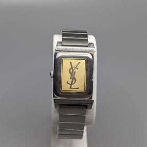 May include: A rectangular wristwatch with a silver-tone metal band and case. The watch face is gold with a black logo. The watch is displayed on a white stand against a gray background.