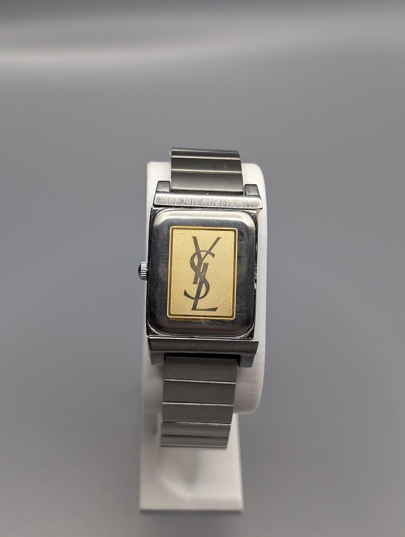 May include: A rectangular wristwatch with a silver-tone metal band and case. The watch face is gold with a black logo. The watch is displayed on a white stand against a gray background.