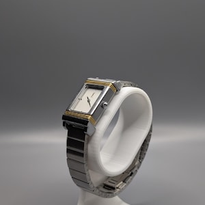 May include: A rectangular wristwatch with a silver-tone metal band and case, accented with gold-tone trim. The watch face is white with black hands and markers. The watch is displayed on a white stand against a gray background.