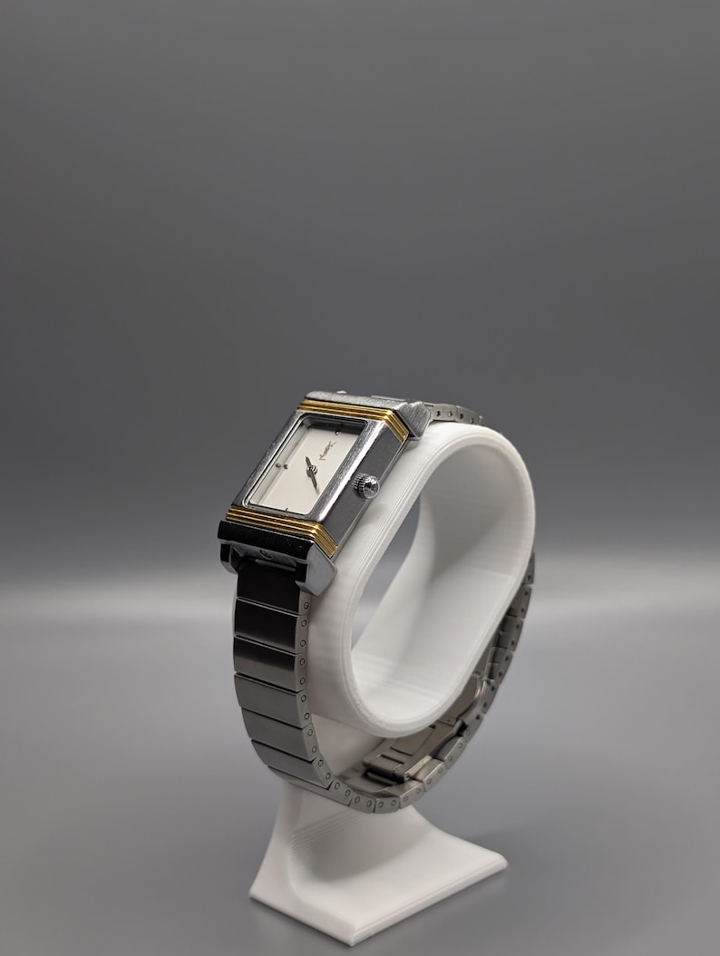 May include: A rectangular wristwatch with a silver-tone metal band and case, accented with gold-tone trim. The watch face is white with black hands and markers. The watch is displayed on a white stand against a gray background.