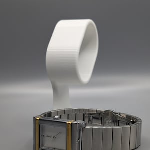 May include: A silver wristwatch with a rectangular face and gold-colored accents. The watch has a silver band and is displayed on a white watch stand. The watch face has a minimalist design.