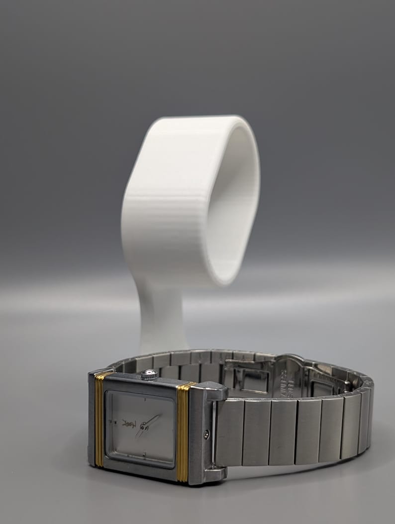 May include: A silver wristwatch with a rectangular face and gold-colored accents. The watch has a silver band and is displayed on a white watch stand. The watch face has a minimalist design.