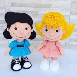 Op de afbeelding: Twee pluche poppen met gezichten in cartoonstijl. De linkerpop heeft zwart haar, een blauwe jurk met twee zwarte horizontale lijnen en zwarte sneakers met witte neuzen. De rechterpop heeft geel krullend haar, een roze jurk en witte sneakers.