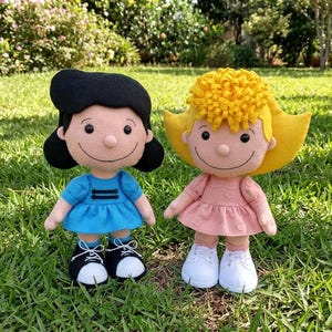 Op de afbeelding: Twee pluche poppen gemaakt van vilt. De linkerpop heeft zwart haar, een blauwe jurk met twee zwarte horizontale balken en zwart-witte sneakers. De rechterpop heeft geel haar, een roze jurk met een stippenpatroon en witte sneakers.