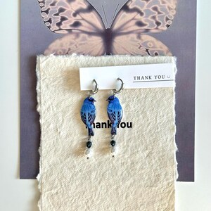 Peut inclure: Boucles d'oreilles bleues en forme d'oiseaux avec des anneaux argentés et des perles pendantes blanches et noires. Elles sont présentées sur une carte blanc cassé texturée avec l'inscription "THANK YOU". Un motif de papillon est en arrière-plan.