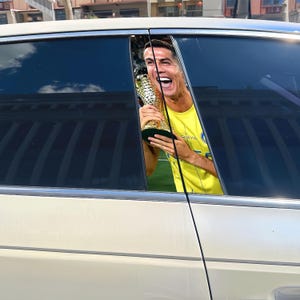 Pode incluir: Um carro branco com um autocolante na janela com um jogador de futebol sorrindo segurando um trof&eacute;u. O jogador est&aacute; vestindo uma camisa amarela. O carro est&aacute; estacionado do lado de fora.