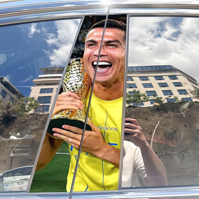 Pode incluir: Fotografia de um homem sorrindo segurando um trof&eacute;u dourado, vestindo uma camisa amarela com a palavra "ROYA" vis&iacute;vel. A imagem &eacute; emoldurada por uma janela de carro, com reflexos de edif&iacute;cios e c&eacute;u.
