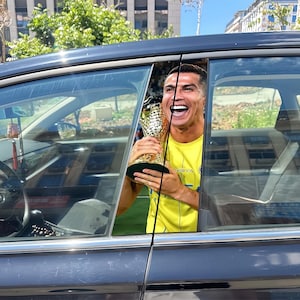 Pode incluir: Uma pessoa segurando um trof&eacute;u de ouro, sorrindo amplamente, vis&iacute;vel atrav&eacute;s da janela de um carro. A pessoa est&aacute; vestindo uma camisa amarela com a palavra "ROYA" impressa. O carro &eacute; preto, e o fundo inclui edif&iacute;cios e vegeta&ccedil;&atilde;o.